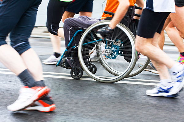 Fotografia przedstawia uliczny bieg. Widoczne są nogi biegaczy. Jeden z nich pcha wózek z osobą niepełnosprawną. Zdjęcie pochodzi z fot. Obrazy licencjonowane przez Depositphotos.com/Drukarnia Chroma.