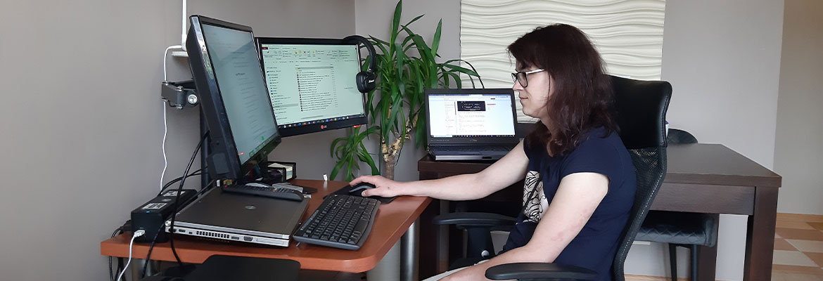 Fotografia przedstawia dziewczynę siedzącą przy biurku z komputerem. Widoczne są dwa monitory, klawiatura i urządzenie wielofunkcyjne. W tle, na stoliku, stoi laptop. Zdjęcie pochodzi z archiwum prywatnego.