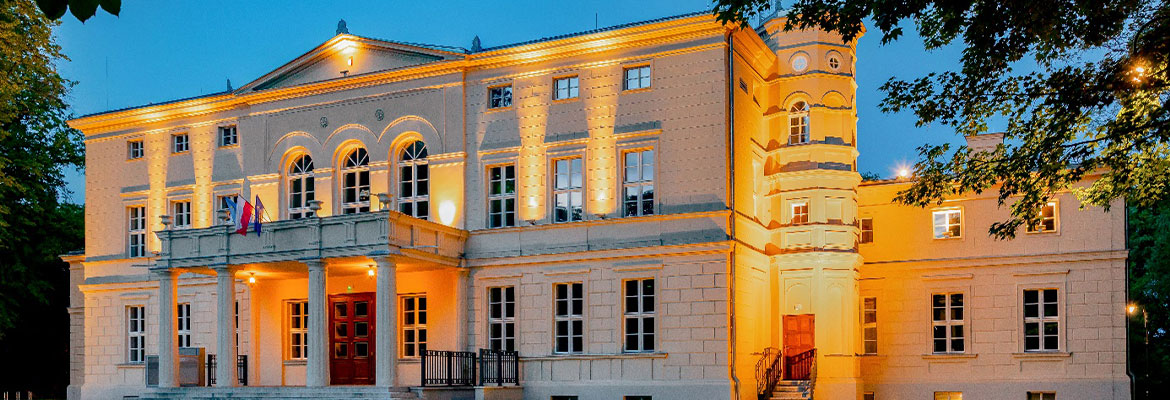 Fotografia przedstawia odnowiony, elegancko podświetlony pałac w Rakoniewicach. Budynek ma białą elewację, wsparty na kolumnach taras nad wejściem głównym czy niewysoką wieżę. Zdjęcie pochodzi z archiwum beneficjenta.