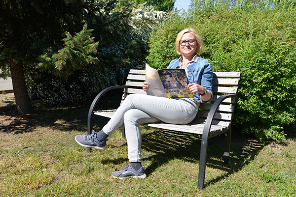 Fotografia przedstawia Lidię Węsierską-Chyc, kobietę, która siedzi na ławce w słonecznej pogodzie i czyta gazetę. Autorem zdjęcia jest Dominik Wójcik.