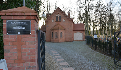 Fotografia wykonana z bramy, przedstawia w tle odnowioną kaplicę cmentarną z czerwonej cegły. Po prawej stronie widoczne są nagrobki z cmentarza. Zdjęcie pochodzi z archiwum beneficjenta.