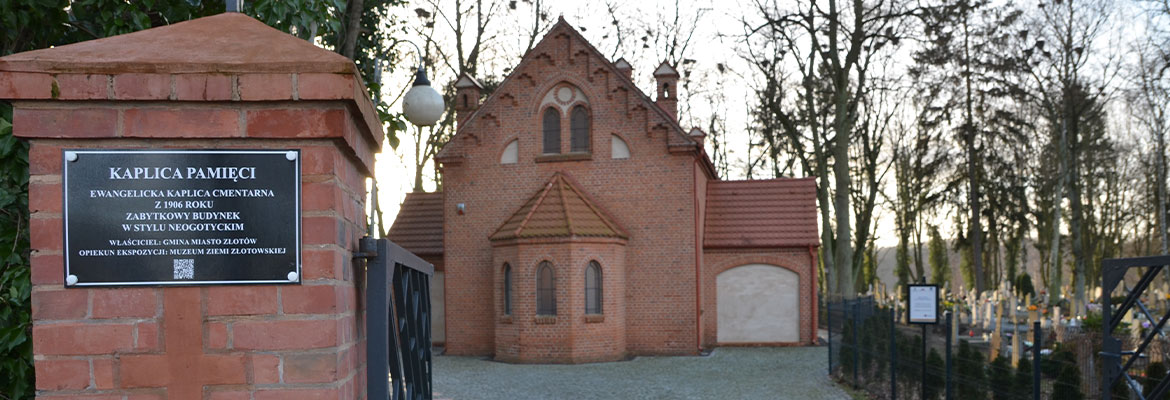 Fotografia wykonana z bramy, przedstawia w tle odnowioną kaplicę cmentarną z czerwonej cegły. Po prawej stronie widoczne są nagrobki z cmentarza. Zdjęcie pochodzi z archiwum beneficjenta.