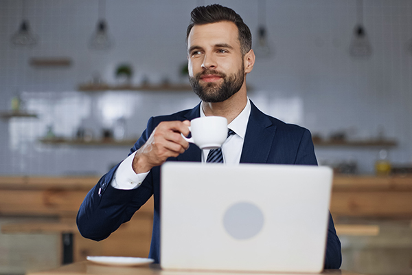 Fotografia przedstawia młodego biznesmena w garniturze, który siedzi przy laptopie z filiżanką kawy w ręku. Zdjęcie pochodzi z Obrazy licencjonowane przez Depositphotos.com/Drukarnia Chroma.