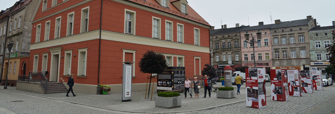 Fotografia przedstawia odnowiony budynek o czerwonej elewacji i spadzistym dachem. Przed nim biegnie chodnik, po którym poruszają się piesi. Autorem zdjęcia jest Dominik Wójcik.