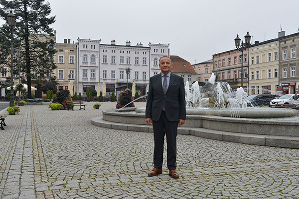 Fotografia przedstawia Piotra Psikusa, burmistrza Kępna. Mężczyzna w garniturze stoi na płycie rynku. Za nim widoczna jest fontanna i okoliczne kamienice. Autorem zdjęcia jest Dominik Wójcik.