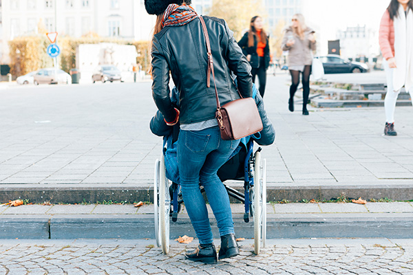 Fotografia przedstawia kobietę, która stara się wepchnąć wózek inwalidzki, na którym siedzi inna osoba, pod wysoki krawężnik. Zdjęcie pochodzi z  Obrazy licencjonowane przez Depositphotos.com/Drukarnia Chroma.