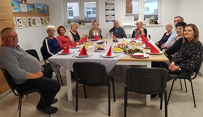 Fotografia przedstawia grupę seniorów, wraz z opiekunem, którzy siedzą przy zastawionym w różne dania, kwadratowym stole. Zdjęcie pochodzi z archiwum beneficjenta.