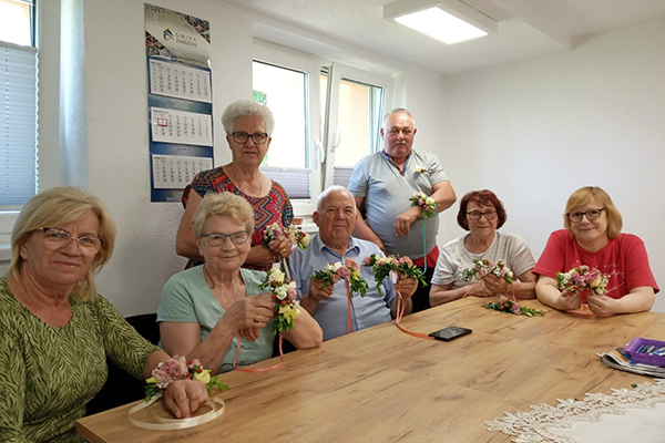 Fotografia przedstawia siedmioro seniorów. Pięcioro siedzi przy stole, dwóch kolejnych stoi za nimi. Wszyscy prezentują wiązanki kwiatów wykonane podczas zajęć florystycznych. Zdjęcie pochodzi z archiwum beneficjenta.