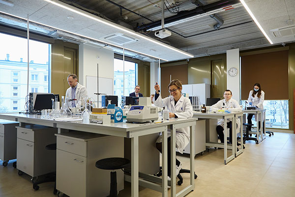Na zdjęciu wykonanym w przestronnej sali widzimy kilka osób w ochronnych białych fartuchach, które wykonują doświadczenia na nowoczesnym sprzęcie laboratoryjnym. Zdjęcie pochodzi z archiwum beneficjenta. 