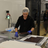 Fotografia przedstawia halę produkcyjną. Widoczne są maszyny i trzy kobiety, które w rękawiczkach, czepkach i słuchawkach, pracują przy czekoladzie. Autorem zdjęcia jest Dominik Wójcik.