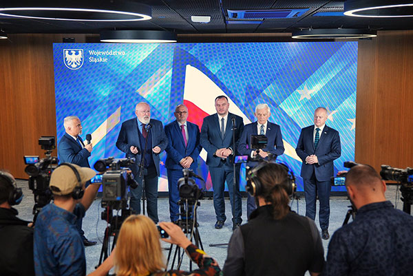 Na zdjęciu widzimy konferencję prasową. Przed kamerami stoi pięciu mężczyzn w garniturach. Obok nich znajduje się inny mężczyzna z mikrofonem. Za nimi wyświetlone jest niebieskie tło z flagą polski. U dołu zdjęcia widoczni są dziennikarze. Zdjęcie pochodzi z archiwum Urzędu Marszałkowskiego Województwa Śląskiego.