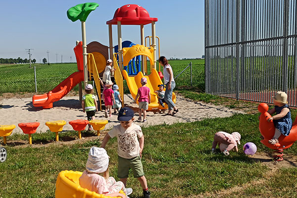 Zdjęcie zrobiono na placu zabaw. Widać na nim kilkanaścioro bawiących się dzieci oraz opiekunkę, która czuwa przy zjeżdżalni. Zdjęcie pochodzi z archiwum beneficjenta.