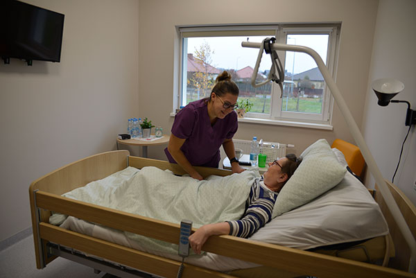 Fotografia przedstawia uśmiechniętą opiekunkę w fioletowym fartuchu, która pomaga ułożyć się w łóżku równie uśmiechniętej pacjentce. Autorem zdjęcia jest Dominik Wójcik.