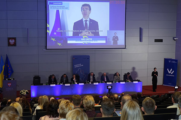 Zdjęcie wykonane z poziomu publiczności ukazuje uczestników konferencji siedzących za stołem oraz mężczyznę, którego wystąpienie wyświetlane jest na ekranie. Zdjęcie pochodzi z archiwum Urzędu Marszałkowskiego Województwa Wielkopolskiego.