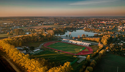Zdjęcie z lotu ptaka przedstawia miejski kwartał. Na pierwszym planie widoczny jest stadion z trybunami, za nim jezioro. Dalej widzimy miejskie zabudowania. Autorem fotografii jest Jarosław Nowicki.