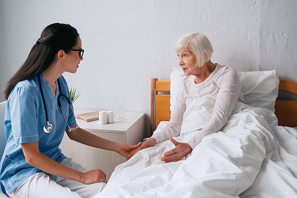 Na fotografii widzimy starszą kobietę leżącą w łóżku, która rozmawia z młodą lekarką. Zdjęcie pochodzi z Obrazy licencjonowane przez Depositphotos.com/Drukarnia Chroma.