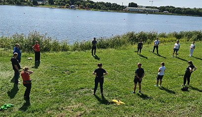 Na fotografii, na zielonej polanie, u brzegu rzeki, ćwiczy z instruktorem kilkanaście osób. Zdjęcie pochodzi z archiwum beneficjenta.