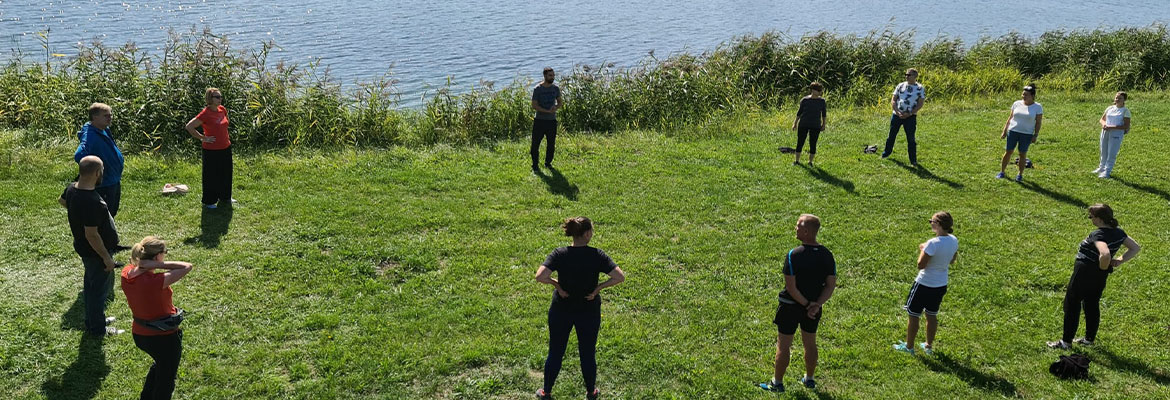 Na fotografii, na zielonej polanie, u brzegu rzeki, ćwiczy z instruktorem kilkanaście osób. Zdjęcie pochodzi z archiwum beneficjenta.