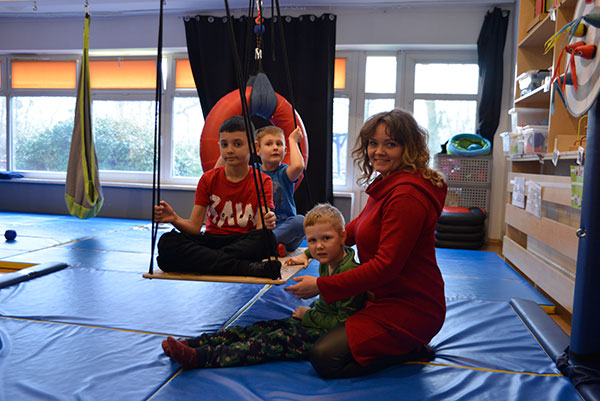Fotografia przedstawia uśmiechniętą kobietę, która na sali gimnastycznej ćwiczy z trójką dzieci. Dwoje z nich siedzi na specjalnej huśtawce. Autorem zdjęcia jest Dominik Wójcik.