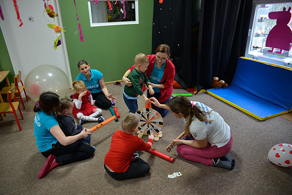 Zdjęcie ukazuje muzykoterapię. Cztery opiekunki i czwórka dzieci siedzą w kole na dywanie. Jedna z opiekunek gra na ukulele. Dzieci losują piosenki za pomocą specjalnej ruletki. Autorem zdjęcia jest Dominik Wójcik.
