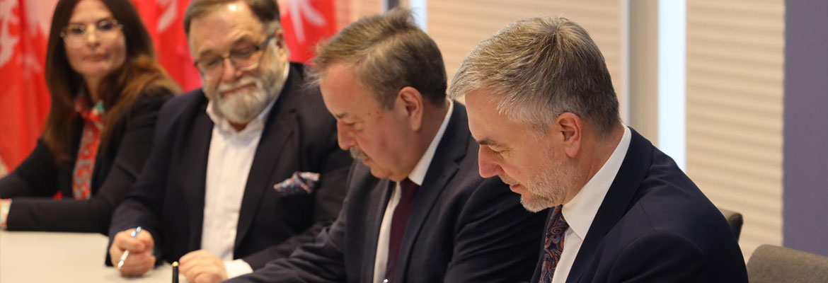  Na zdjęciu widzimy trzech mężczyzn w garniturach i kobietę. Wszyscy siedzą przy stole i podpisują umowę. Zdjęcie pochodzi z archiwum Urzędu Marszałkowskiego Województwa Wielkopolskiego.