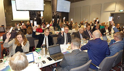 Na zdjęciu widzimy kilkadziesiąt osób, obradujących przy dwóch długich stołach. Wszyscy wyciągają w górę rękę z kartą do głosowania. Zdjęcie pochodzi z Urzędu Marszałkowskiego Województwa Wielkopolskiego.