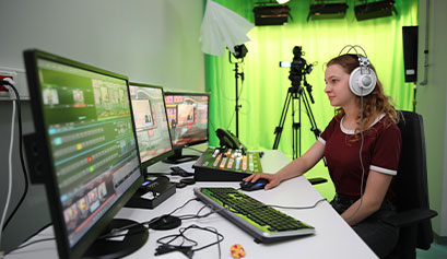 Na zdjęciu widzimy uczennicę ze słuchawkami na głowie, która siedzi przy stanowisku komputerowym złożonym z trzech monitorów, w studiu filmowo-fotograficzny. Fotografia pochodzi z archiwum beneficjenta.