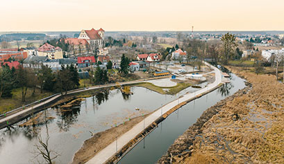 Na pierwszym planie zdjęcia widzimy rzekę ze ścieżkami biegnącymi wzdłuż oraz przystaniami. W tle widać zaś zabudowania miasta. Autorem fotografii jest Jakub Kosmatka.