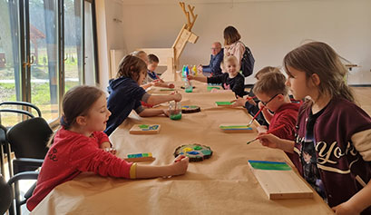 Na zdjęciu widzimy dzieci siedzące wzdłuż podłużnego stołu, malujące na drewnie. Fotografia pochodzi z archiwum beneficjenta.