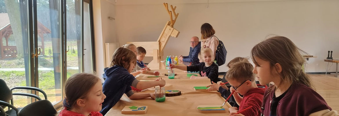 Na zdjęciu widzimy dzieci siedzące wzdłuż podłużnego stołu, malujące na drewnie. Fotografia pochodzi z archiwum beneficjenta.