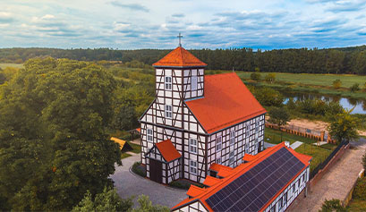 Zdjęcie z lotu ptaka przedstawia niewielki kościół z muru pruskiego, pokryty czerwoną dachówką, położony na brzegu rzeki. Fotografia pochodzi z archiwum beneficjenta.