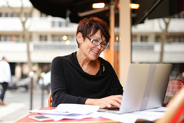 Na zdjęciu widzimy kobietę w średnim wieku, która z uśmiechem na twarzy pracuje przy laptopie. Fotografia pochodzi z adobe stock.