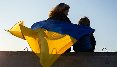 Na zdjęciu widać plecy kobiety i dziecka, którzy siedzą na betonowym murku. Na plecach powiewa im flaga Ukrainy. Fotografia pochodzi z adobe stock.