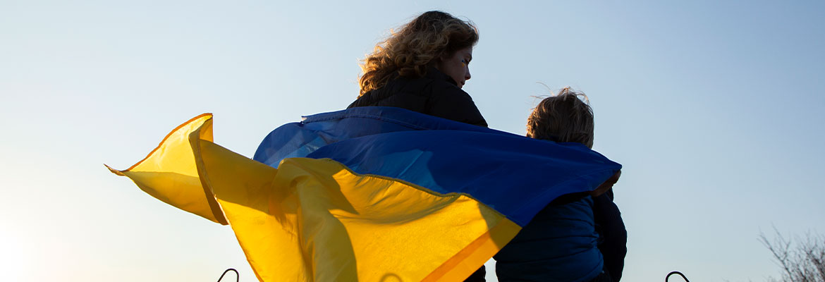 Na zdjęciu widać plecy kobiety i dziecka, którzy siedzą na betonowym murku. Na plecach powiewa im flaga Ukrainy. Fotografia pochodzi z adobe stock.