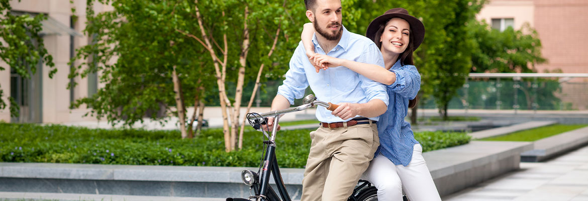 Fotografia przedstawia elegancko ubranych kobietę i mężczyznę, którzy jadą rowerem. Za nimi znajduje się niewielka, zielona przestrzeń. Zdjęcie pochodzi z chroma stock.