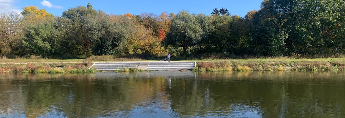 Fotografia wykonana jest z brzegu rzeki. Widoczne jest jej koryto oraz drugi brzeg, na którym stoi rowerzysta. Zdjęcie pochodzi z archiwum beneficjenta.