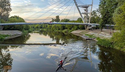Na zdjęciu widoczna jest nowoczesna kładka łącząca dwa zalesione brzegi rzeki. Po wodzie płynie kajakarz. Autorem zdjęcia jest Marcin Dekert.