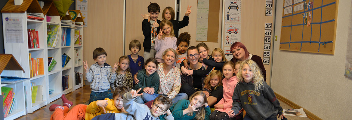 Na zdjęciu widoczna jest grupa dzieciaków z nauczycielkami. Wszyscy siedzą na dywanie w niewielkiej sali. Autorem fotografii jest Dominik Wójcik.