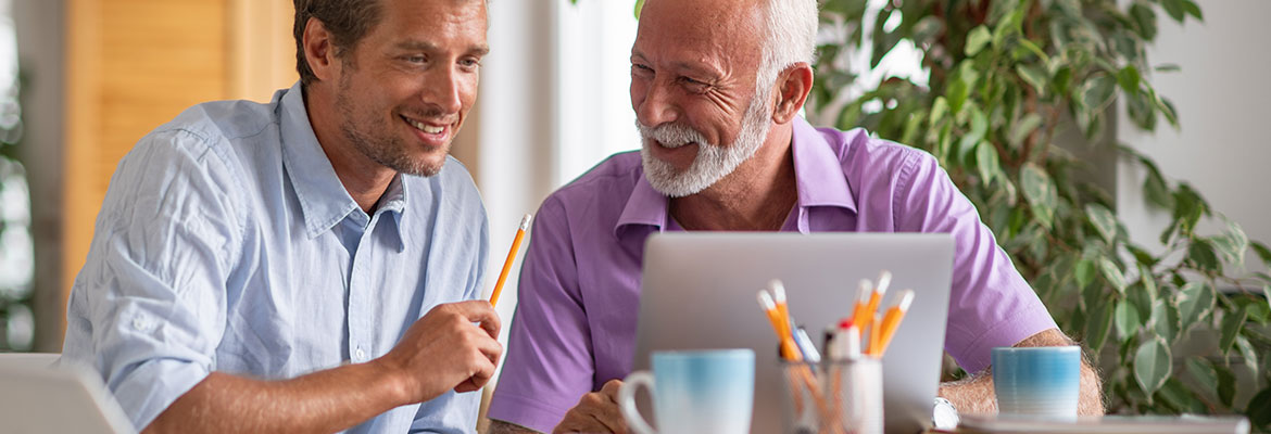 Na zdjęciu znajduje się dwóch uśmiechniętych mężczyzn – jeden młody, drugi starszy. Siedzą przy stole i patrzą w ekran laptopa. Fotografia pochodzi z adobe stock.