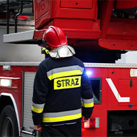 Na fotografii widoczny jest strażak, który stoi plecami do fotografa, na tle wozu strażackiego. Zdjęcie pochodzi z adobe stock.
