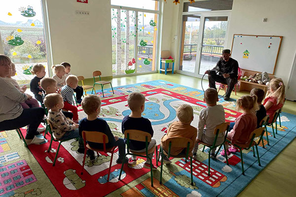 Fotografię wykonano w sali przedszkolnej. Dzieci się dą na krzesełkach ustawionych w kształcie litery L i słuchają swojego gościa – kominiarza. Zdjęcie pochodzi z archiwum beneficjenta.