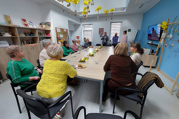 Zdjęcie przedstawia salę w klubie seniora. Uczestnicy siedzą przy stole i słuchają prowadzącego zajęcia. Fotografia pochodzi z archiwum beneficjenta.