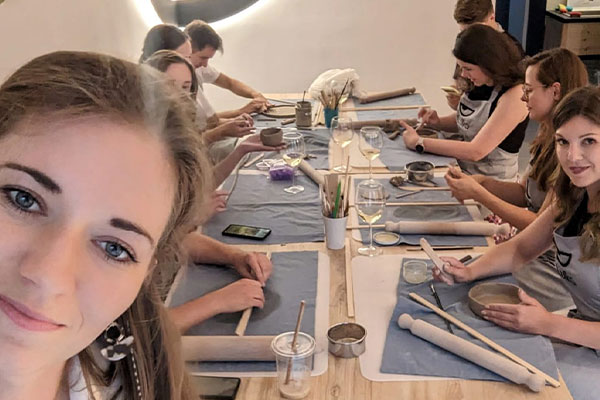 Zdjęcie to tak zwane selfie. Widoczna jest twarz kobiety, która je wykonuje, a w tle siedzące przy stole kolejne osoby, które tworzą ceramikę. Fotografia pochodzi z archiwum prywatnego.