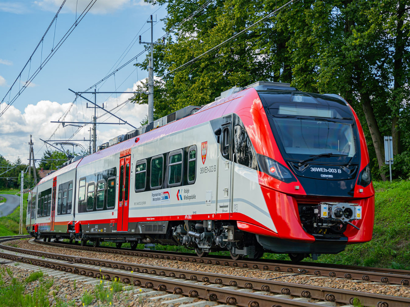 Na zdjęciu widoczny jest pociąg typu „Elf”, w biało-czerwonych barwach, z herbem województwa wielkopolskiego, sunący po torach. Fotografia pochodzi z archiwum Kolei Wielkopolskich.