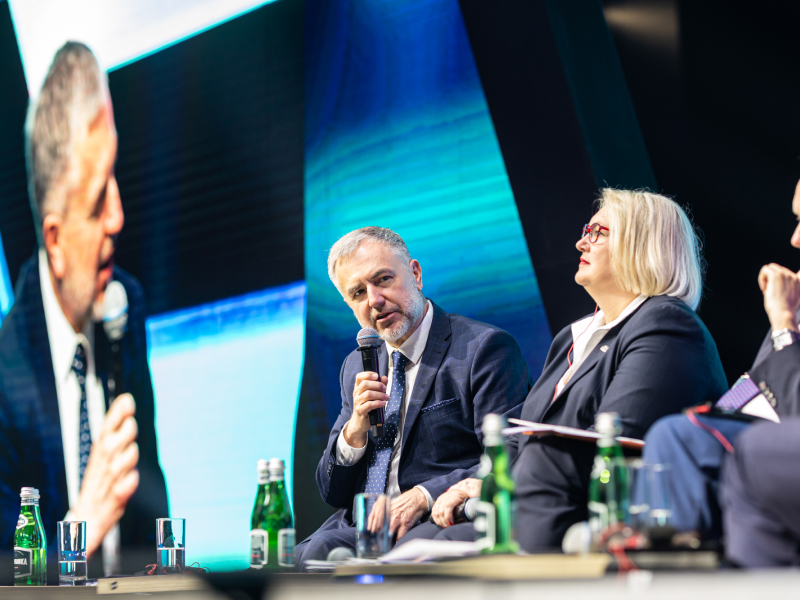 Zdjęcie przedstawia fragment konferencji. Ujęcie ukazuje trzy osoby siedzące na scenie. Jedna z nich zabiera głos. Za nią, w tle, widoczny jest telebim, który przekazuje na żywo obraz ze sceny. Fotografia pochodzi z archiwum Samorządu Województwa Wielkopolskiego.