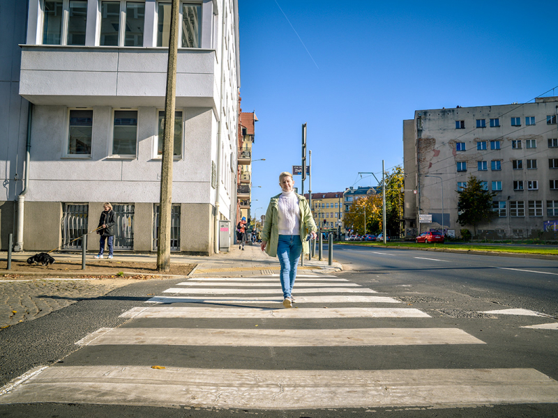 Na zdjęciu widoczna jest kobieta, która przechodzi przez przejście dla pieszych w mieście. Autorem fotografii jest Dominik Wójcik.