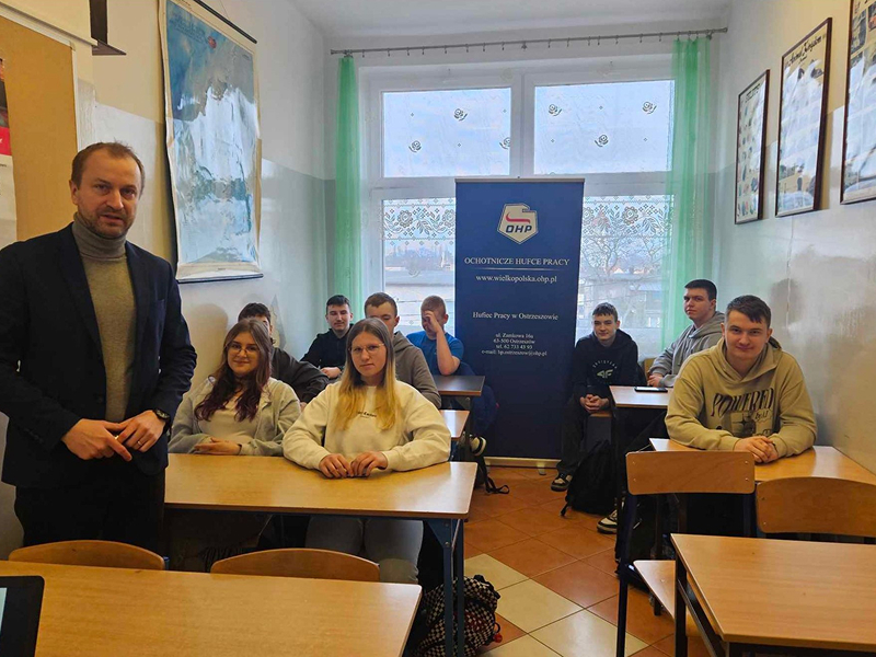 Zdjęcie wykonano w sali lekcyjnej. Na pierwszym planie widoczny jest stojący nauczyciel w ciemnej marynarce. Za nim, w ławkach, siedzą uczniowie. Fotografia pochodzi z archiwum beneficjenta.