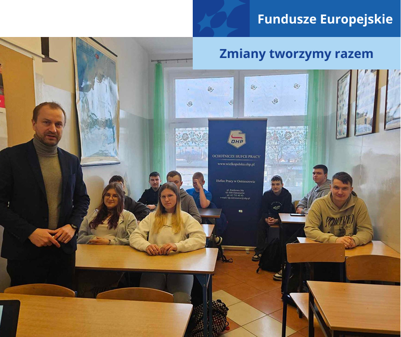 Zdjęcie wykonano w sali lekcyjnej. Na pierwszym planie widoczny jest stojący nauczyciel w ciemnej marynarce. Za nim, w ławkach, siedzą uczniowie. Fotografia pochodzi z archiwum beneficjenta.