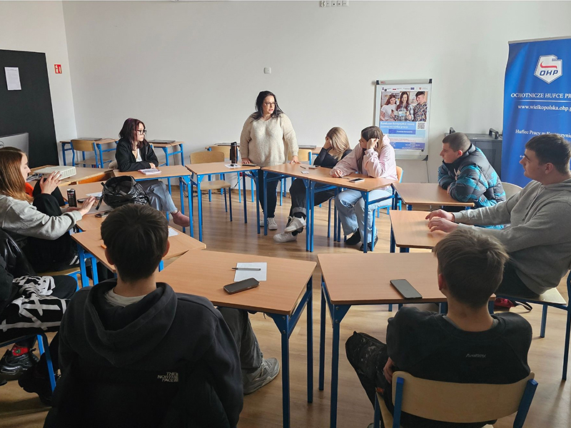 Fotografia przedstawia salę w trakcie zajęć. Przy stołach ustawionych w okrąg siedzą młodzi uczestnicy. Wśród nich jest prowadząca zajęcia kobieta, która stoi. Zdjęcie pochodzi z archiwum beneficjenta.