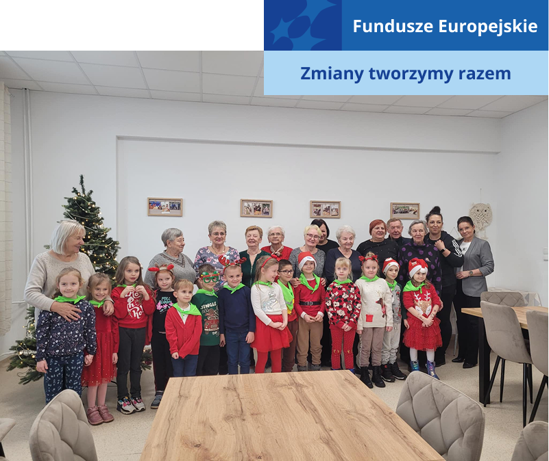 Na zdjęciu widoczna jest grupa seniorów, przed którą stoi grupa małych dzieci. Fotografię wykonano w pomieszczeniu należącym do klubu seniora. Pochodzi z archiwum beneficjenta.